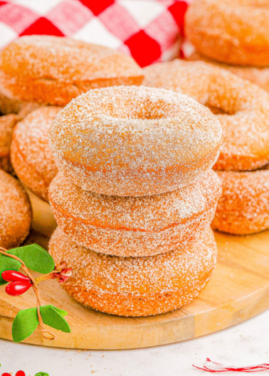 A stack of sugar-coated donuts on a wooden board, with a red-and-white checkered cloth and a small holly decoration nearby.