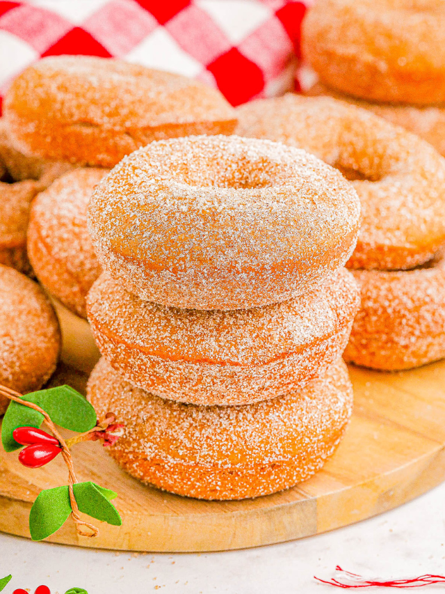 A stack of sugar-coated donuts on a wooden board, with a red-and-white checkered cloth and a small holly decoration nearby.