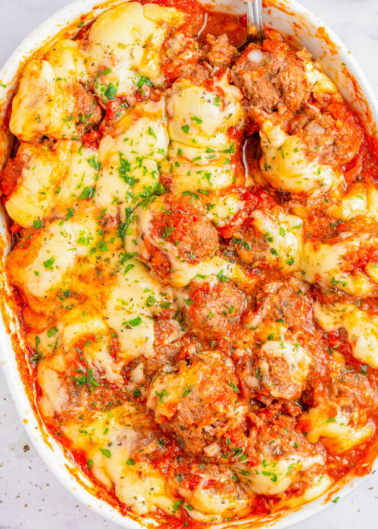 Close-up of a baked dish with meatballs, tomato sauce, melted cheese, and sprinkled herbs in a white oval dish.