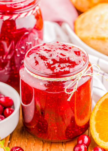 A jar of homemade cranberry sauce sits on a table, surrounded by fresh cranberries and a sliced orange. Another jar and bread are in the background.