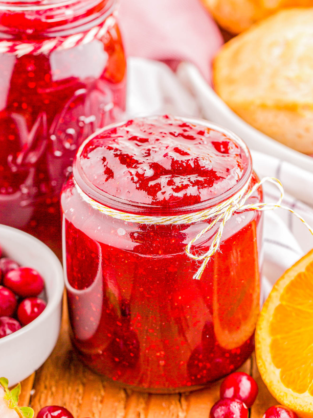 A jar of homemade cranberry sauce sits on a table, surrounded by fresh cranberries and a sliced orange. Another jar and bread are in the background.