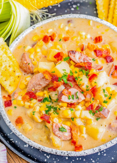 A bowl of creamy corn chowder with sliced sausage, diced potatoes, and garnished with bacon bits and parsley. Corn husks and a corn cob are visible in the background.