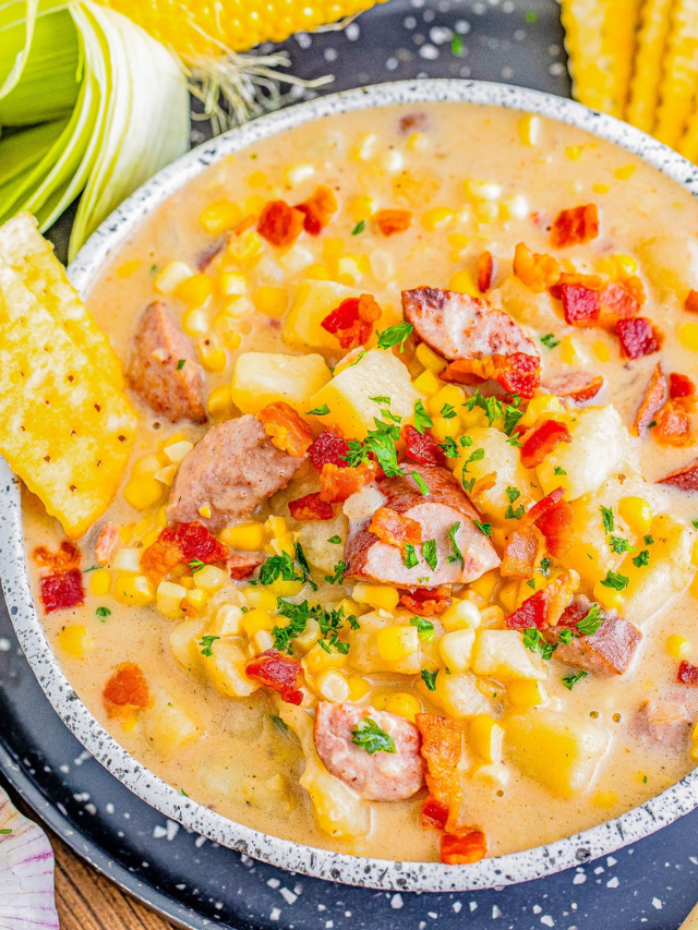 A bowl of creamy corn chowder with sliced sausage, diced potatoes, and garnished with bacon bits and parsley. Corn husks and a corn cob are visible in the background.