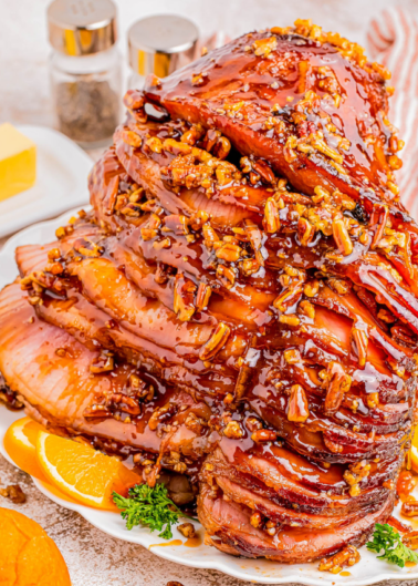 A glazed, sliced ham garnished with orange slices and parsley on a platter. Peppershakers and butter are in the background.