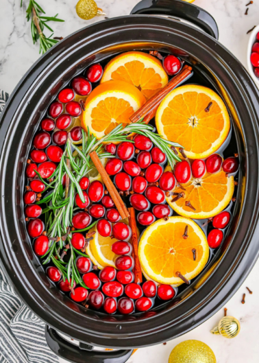 Slow cooker with sliced oranges, cranberries, rosemary, and cinnamon sticks.