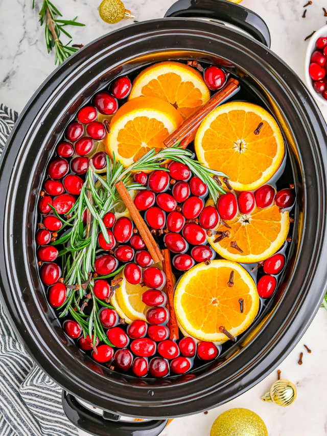 Slow cooker with sliced oranges, cranberries, rosemary, and cinnamon sticks.