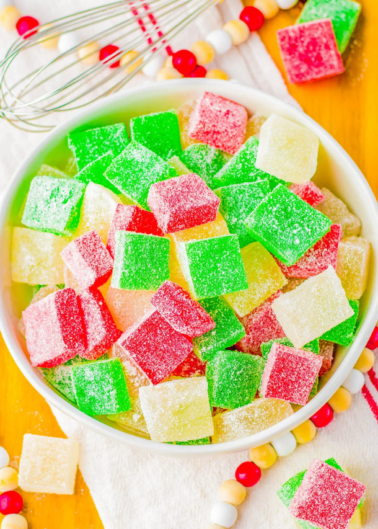 A bowl of colorful sugar-coated gummy cubes in green, red, and yellow. A whisk and beaded decorations are visible in the background.