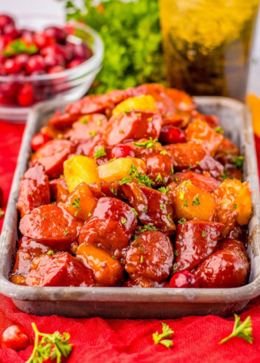 A dish of glazed sausages with pineapples and cranberries in a metal tray, garnished with herbs.