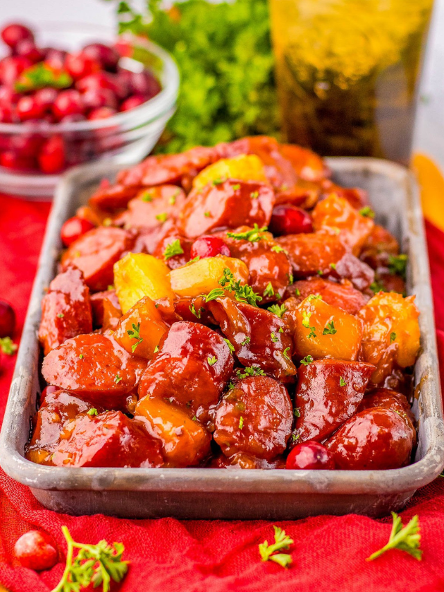 A dish of glazed sausages with pineapples and cranberries in a metal tray, garnished with herbs.