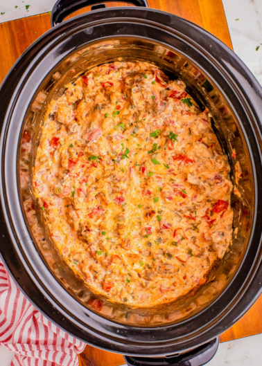 A slow cooker filled with a creamy dip, featuring visible chopped tomatoes and herbs, placed on a wooden table.