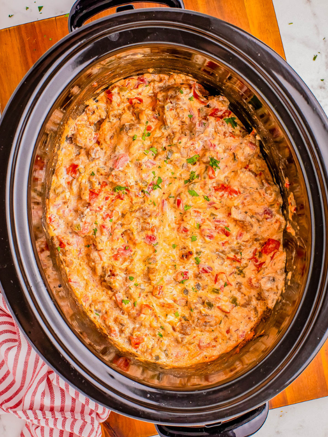 A slow cooker filled with a creamy dip, featuring visible chopped tomatoes and herbs, placed on a wooden table.