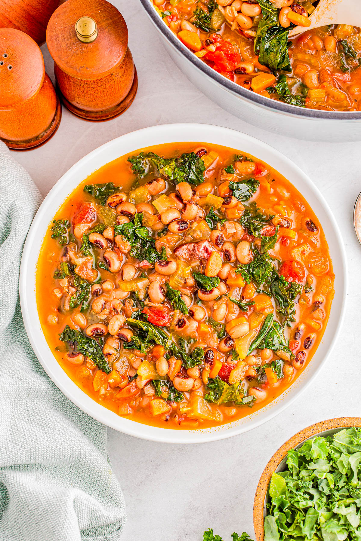 A bowl of bean soup filled with kale, tomatoes, and vegetables on a white surface, next to a green cloth and wooden salt and pepper shakers.