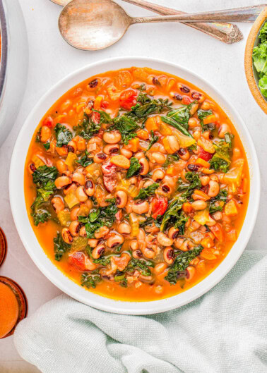 A bowl of soup with black-eyed peas, kale, tomatoes, and visible seasonings, placed on a white surface next to two spoons and a green cloth.