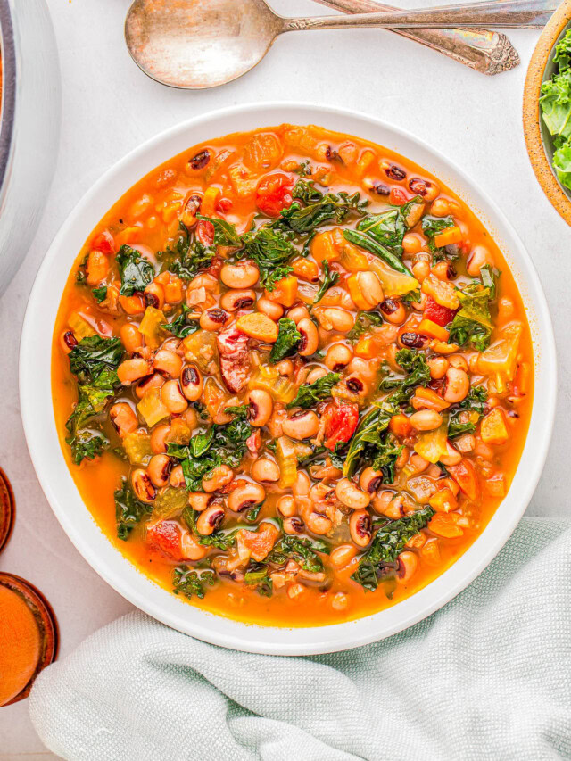 A bowl of soup with black-eyed peas, kale, tomatoes, and visible seasonings, placed on a white surface next to two spoons and a green cloth.