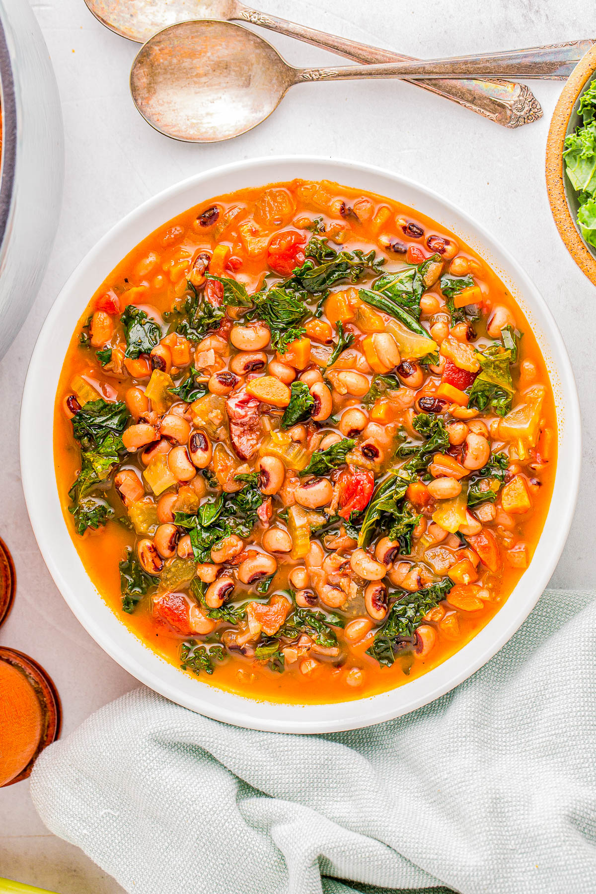 A bowl of soup with black-eyed peas, kale, tomatoes, and visible seasonings, placed on a white surface next to two spoons and a green cloth.