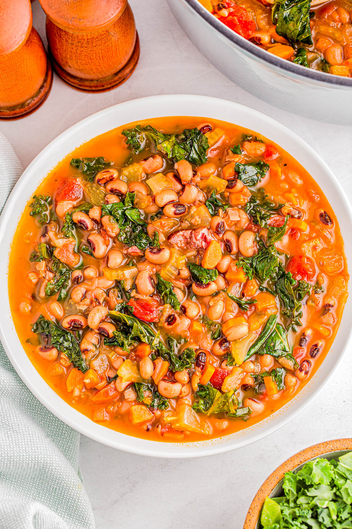 A bowl of stew with black-eyed peas, kale, tomatoes, and vegetables in a tomato-based broth, placed on a white table near pepper shakers.