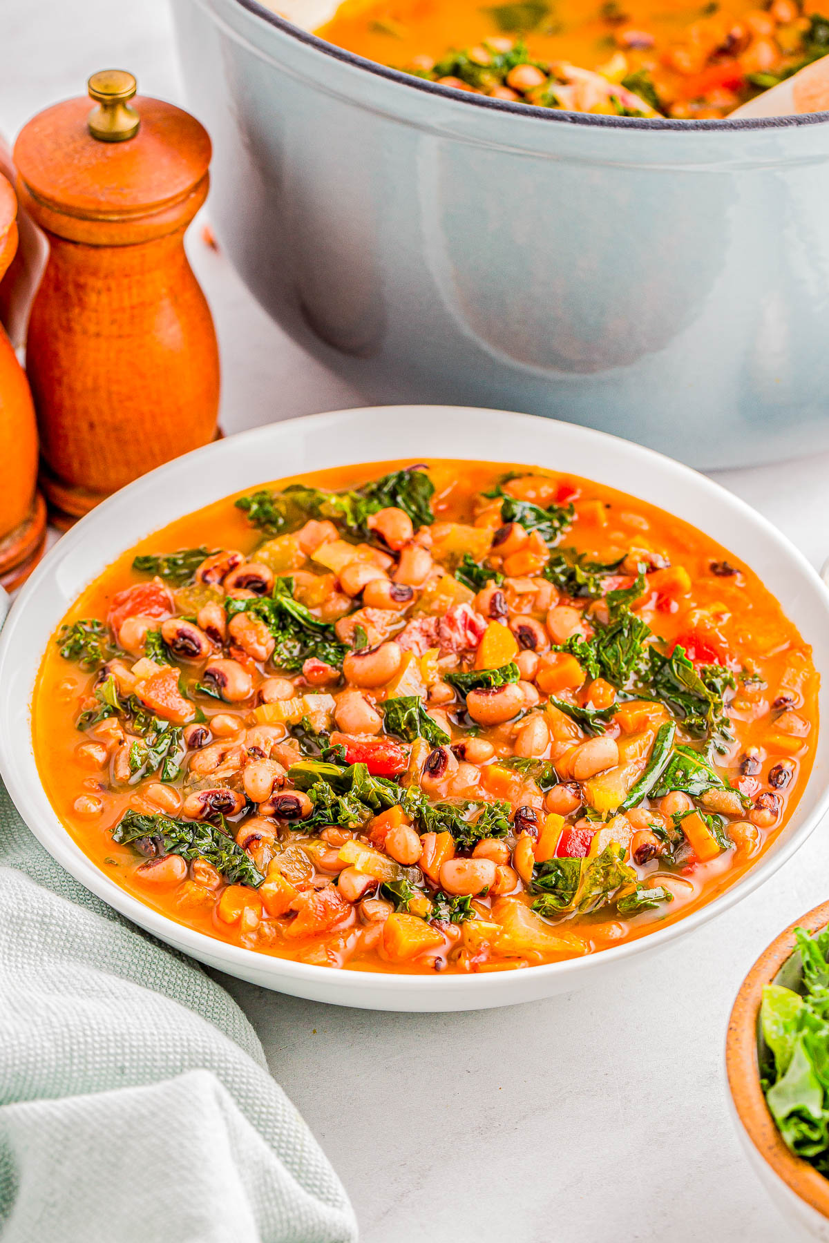 A bowl of vegetable soup with beans, greens, and tomatoes, next to a pot and salt and pepper shakers on a white surface.