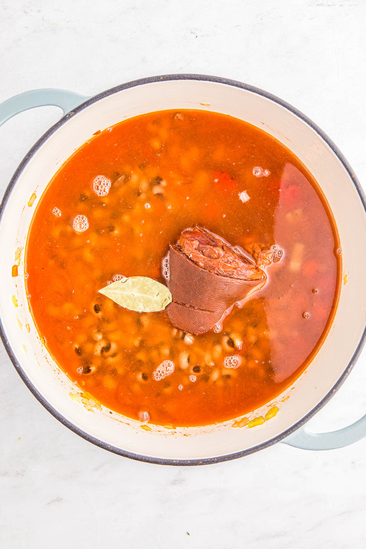 A pot of soup with beans, a bay leaf, and a ham hock on a light-colored surface.