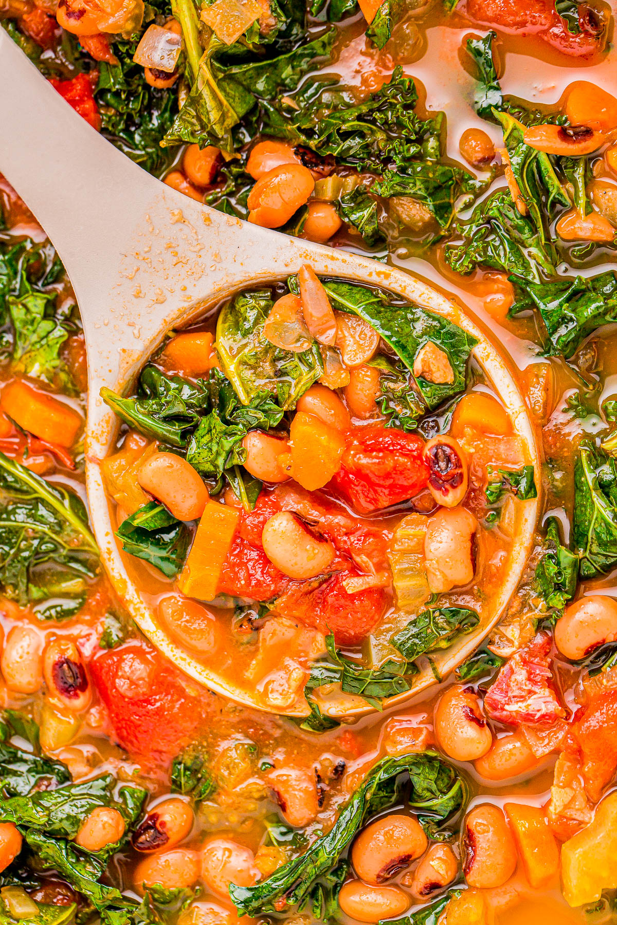 A close-up of a vegetable stew with black-eyed peas, kale, tomatoes, and bell peppers. A white ladle scoops a portion of the colorful mixture.