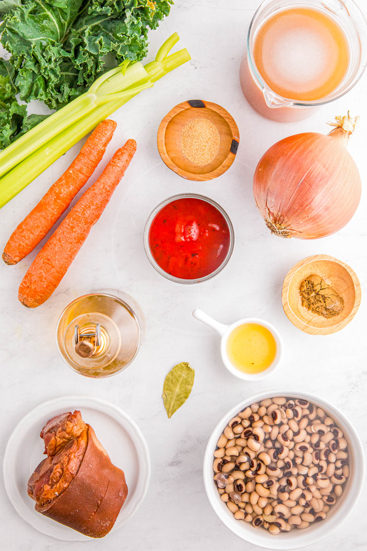 Ingredients for a soup recipe: kale, celery, carrots, onion, broth, tomato paste, seasonings, black-eyed peas, a ham hock, and a bay leaf arranged on a white surface.