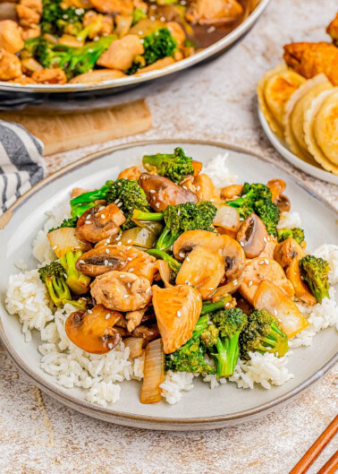 A plate of stir-fried chicken, mushrooms, and broccoli served over white rice. Nearby, there is a plate with dumplings and fried chicken.