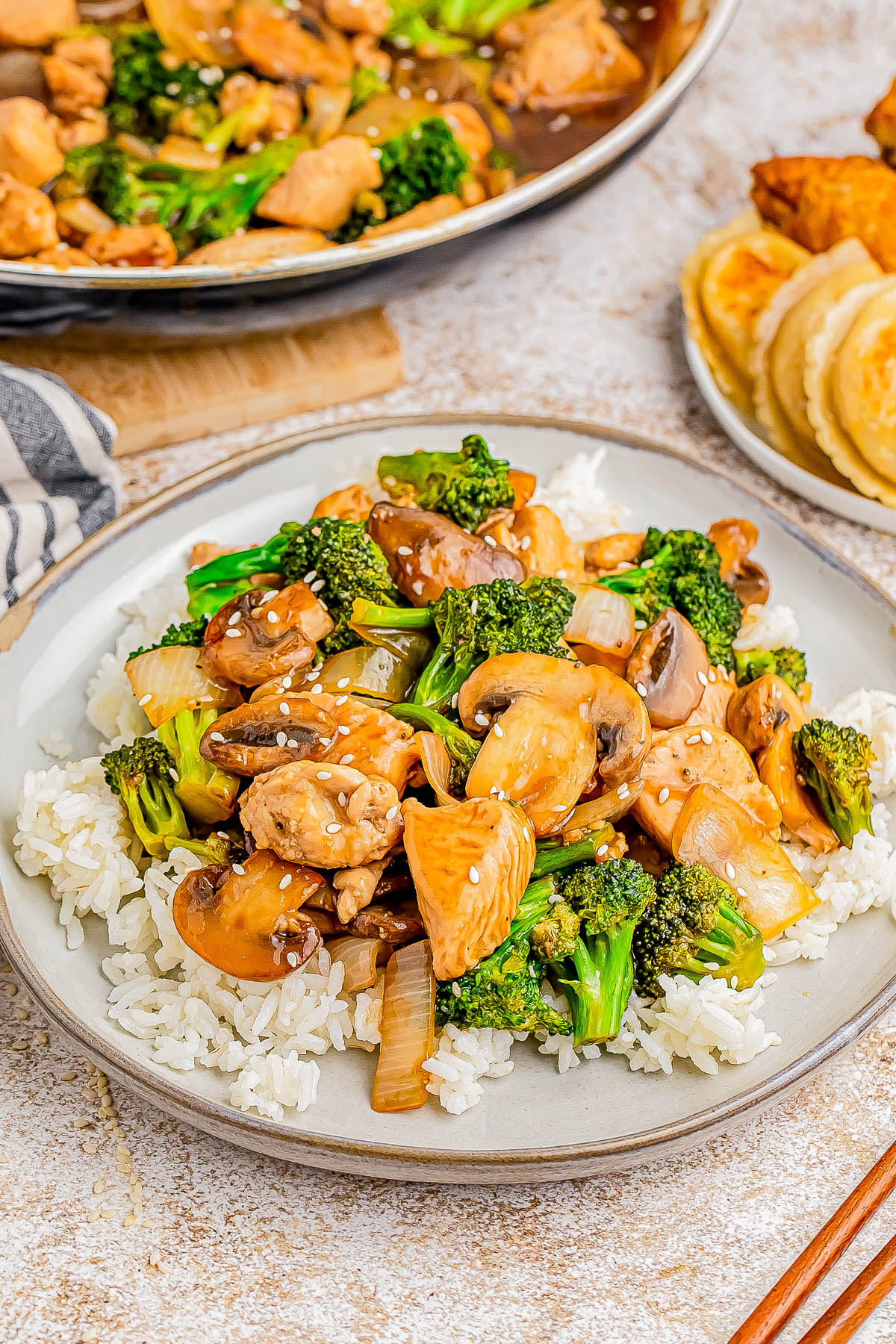 A plate of stir-fried chicken, mushrooms, and broccoli served over white rice. Nearby, there is a plate with dumplings and fried chicken.