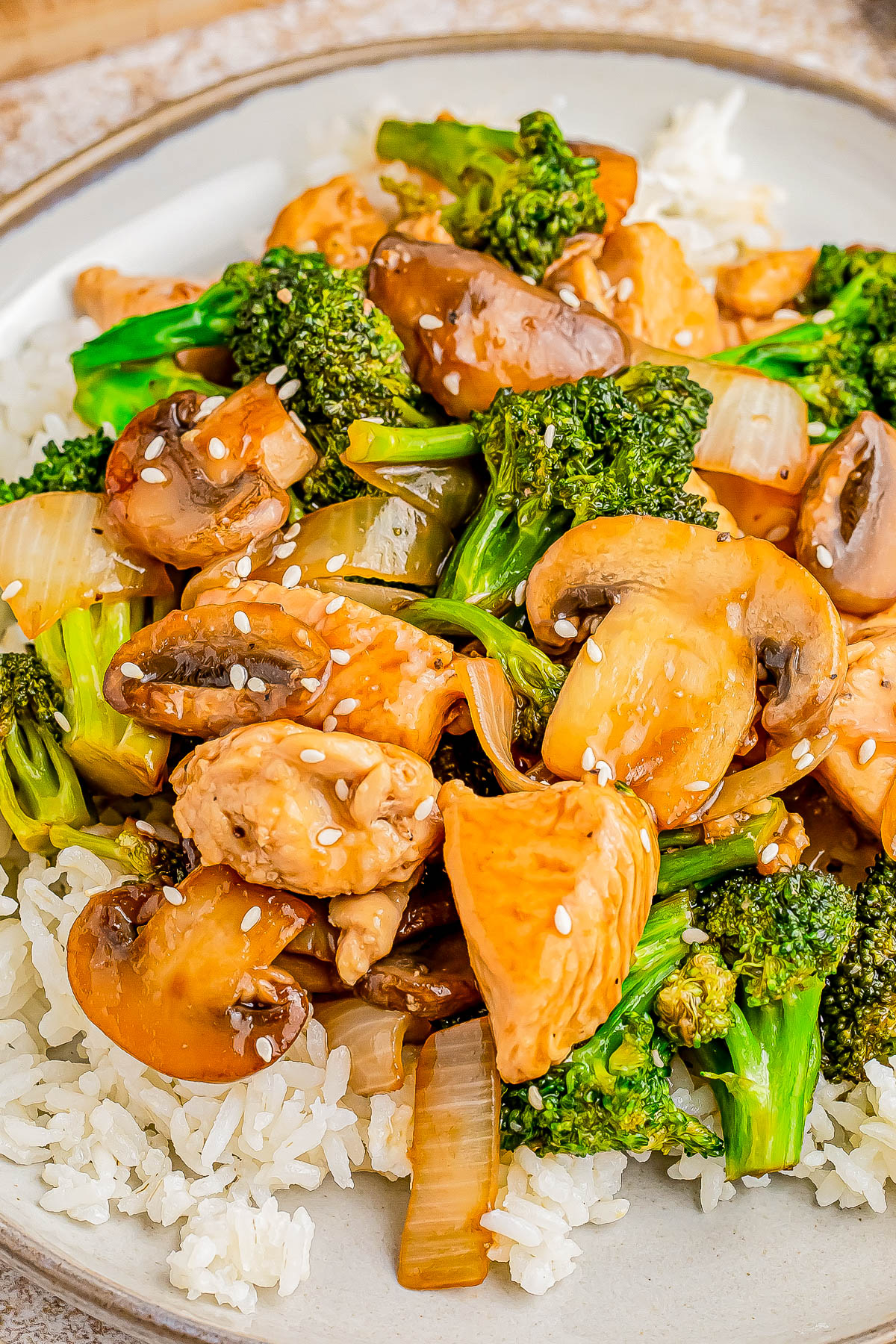 A plate of chicken stir-fry with broccoli, mushrooms, onions, and sesame seeds, served over white rice.