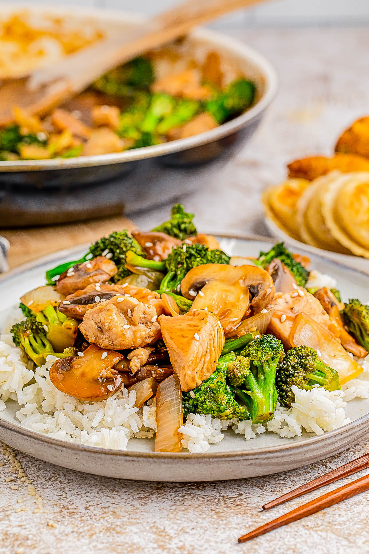 Plate of stir-fried chicken with broccoli and mushrooms on white rice, garnished with sesame seeds, with a pan and dumplings in the background.