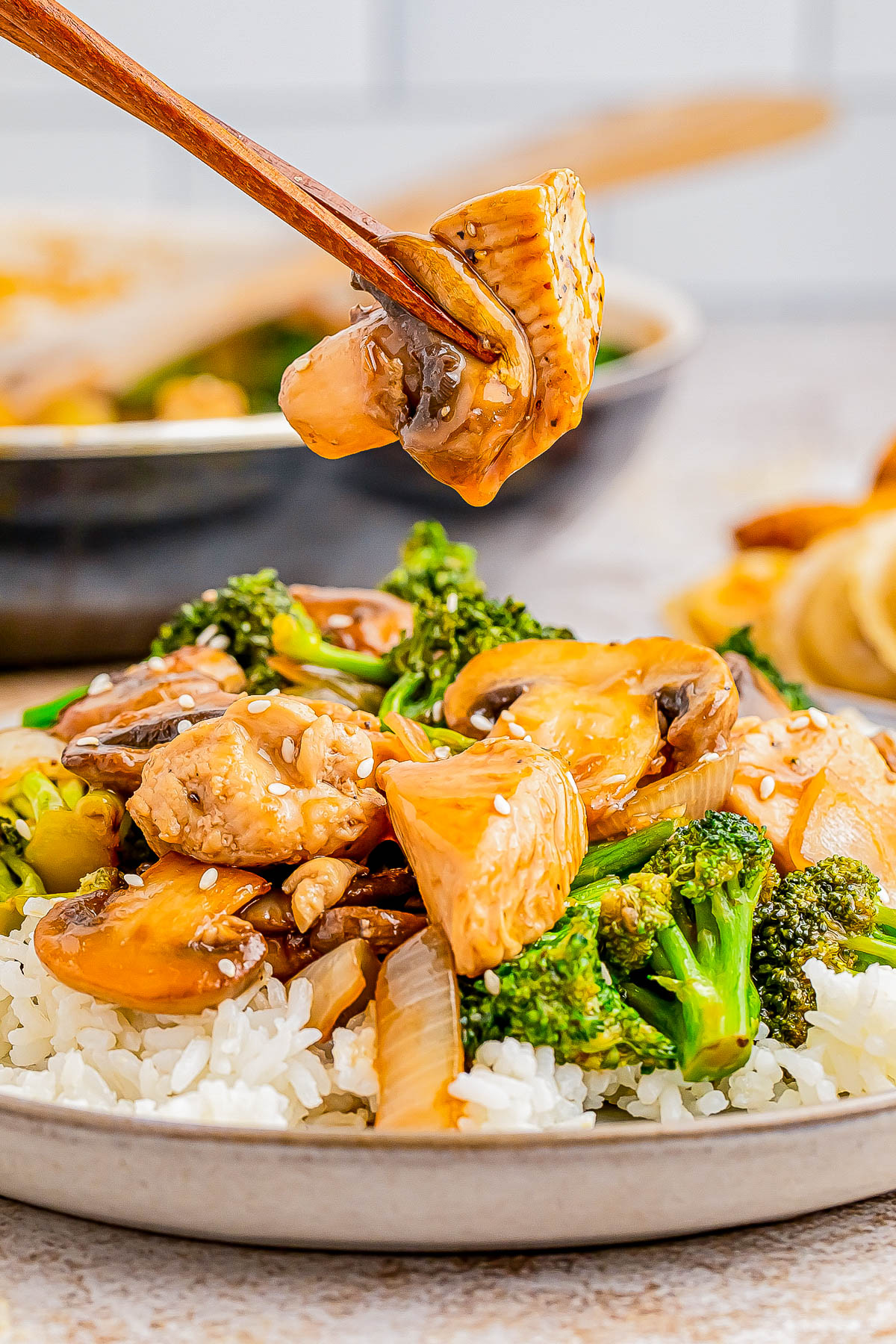 A dish of stir-fried chicken, broccoli, and mushrooms over rice, with chopsticks holding a piece of chicken.