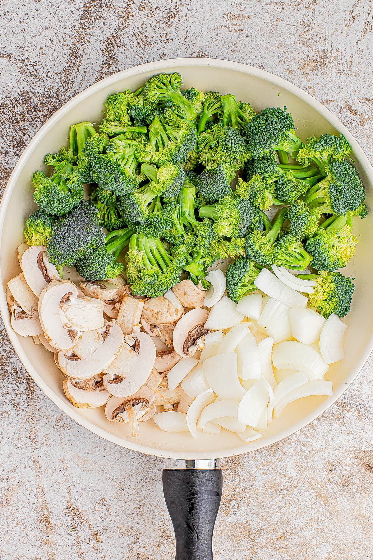 A frying pan with chopped broccoli, sliced mushrooms, and diced onions on a textured surface.