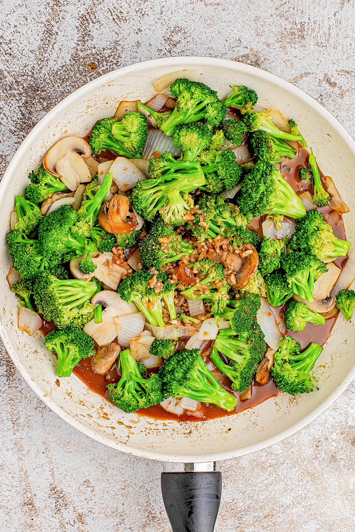 A skillet contains cooked broccoli, onions, and mushrooms in a savory sauce.