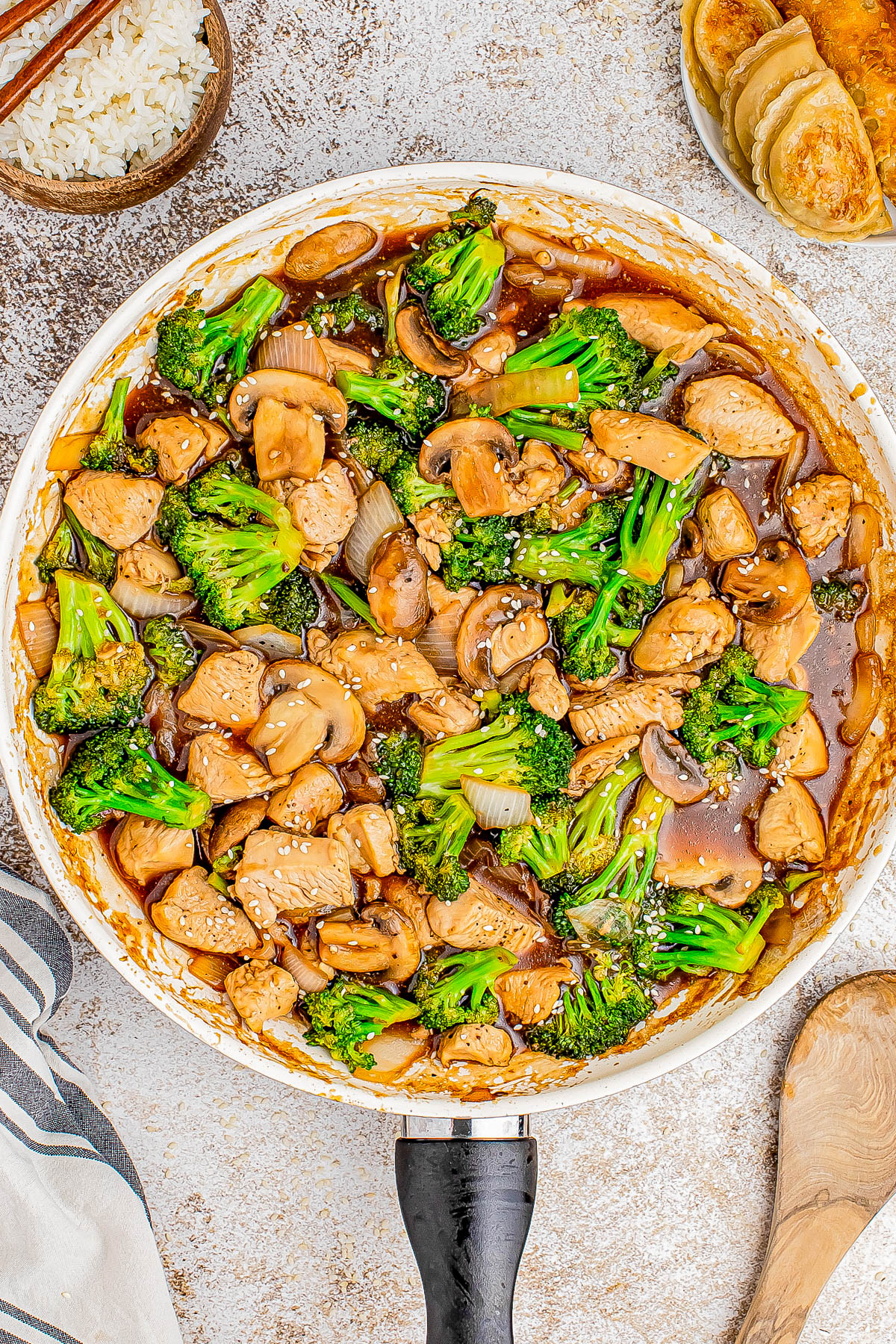 A pan filled with stir-fried chicken, broccoli, mushrooms, and onions in a brown sauce, sprinkled with sesame seeds. Rice and dumplings are on the side.