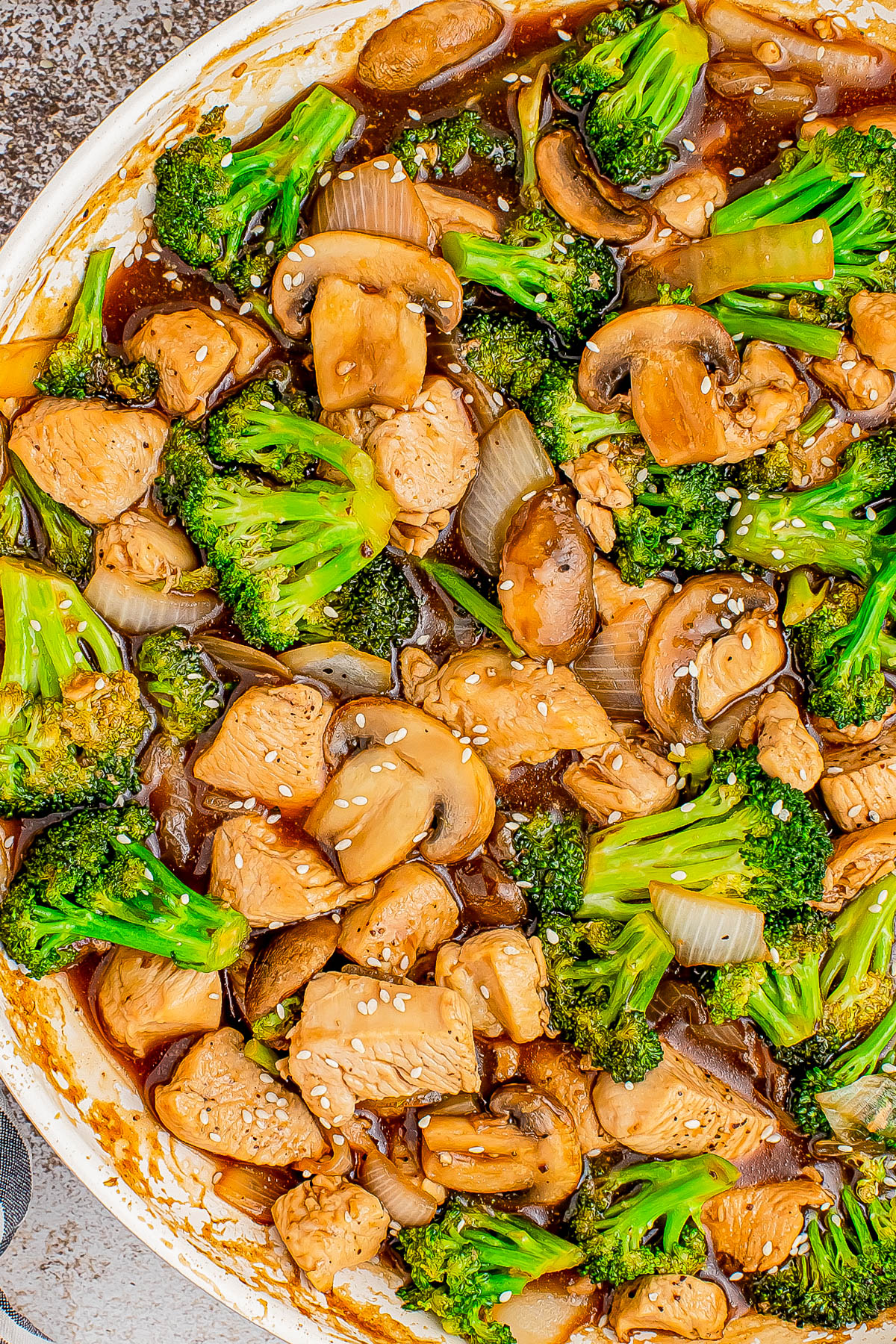 A stir-fry dish with chicken, broccoli, mushrooms, and onions in a brown sauce, garnished with sesame seeds.