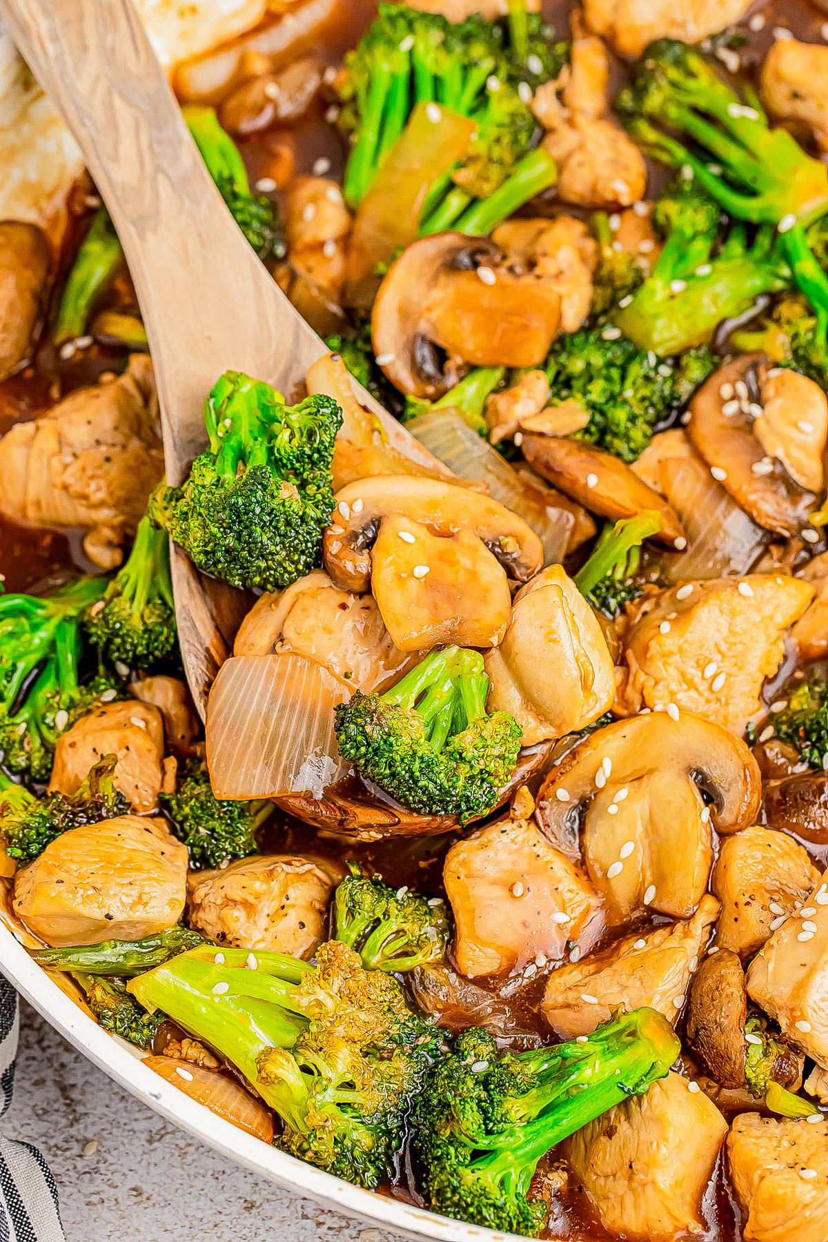 A wooden spoon scoops a stir-fry dish with chicken, broccoli, mushrooms, onions, and sesame seeds in a thick sauce from a white pan.