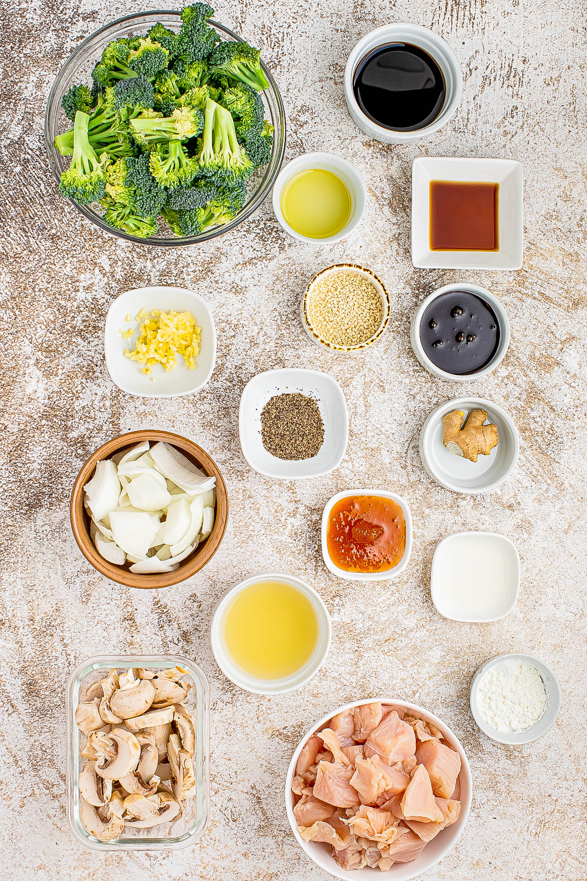 An assortment of ingredients including broccoli, chicken, mushrooms, onions, and various sauces and seasonings arranged in bowls on a textured surface.