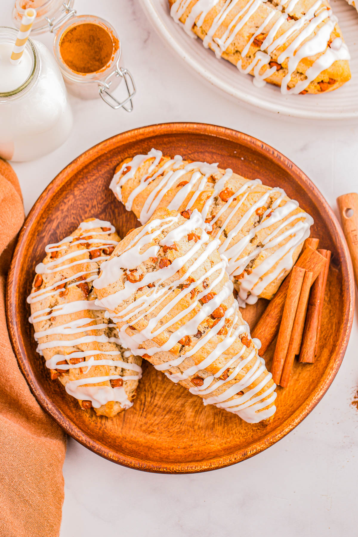 Three iced scones on a wooden plate with cinnamon sticks beside them. A jar of cinnamon and a bottle of milk are nearby.