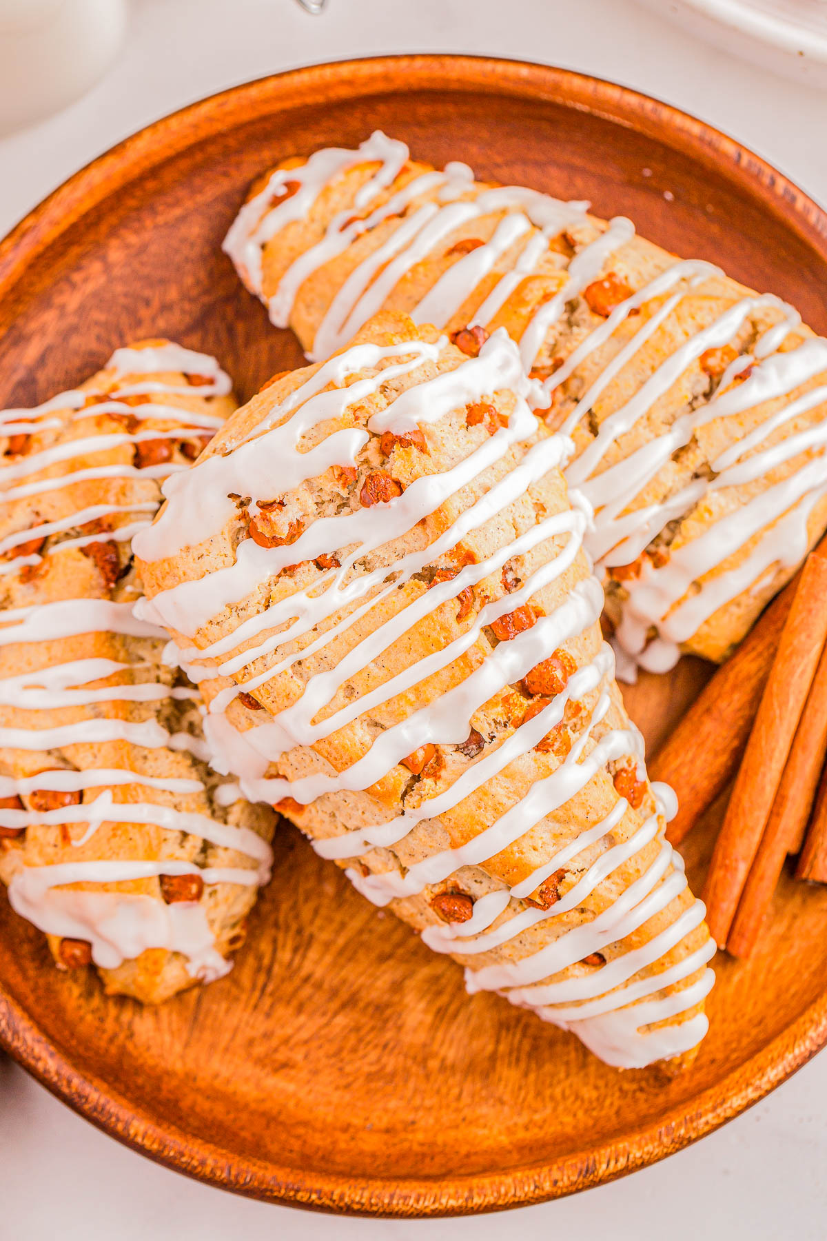 Three scones drizzled with white icing on a wooden plate, accompanied by cinnamon sticks.