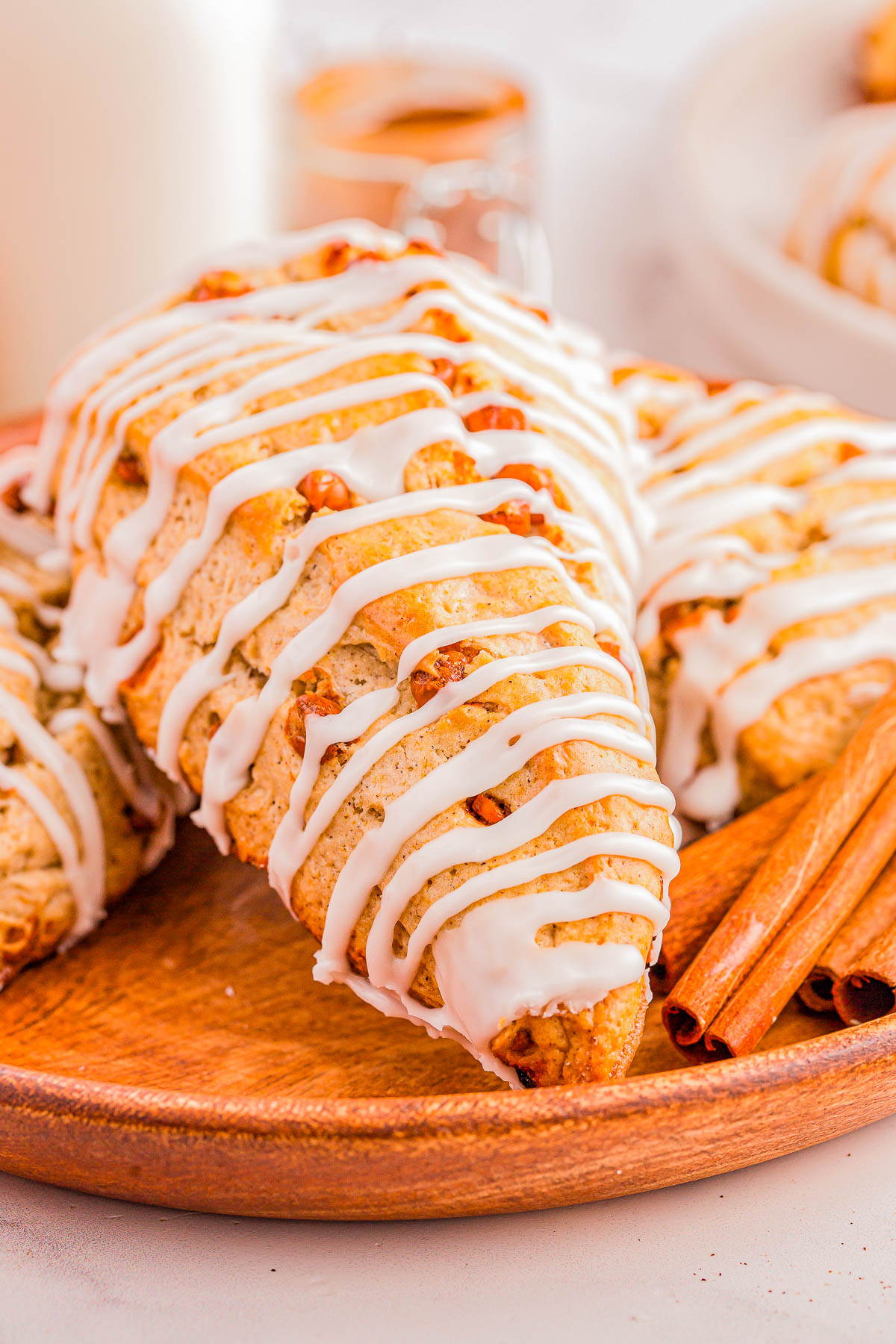 Close-up of a cinnamon scone drizzled with white icing on a wooden plate, with cinnamon sticks beside it.