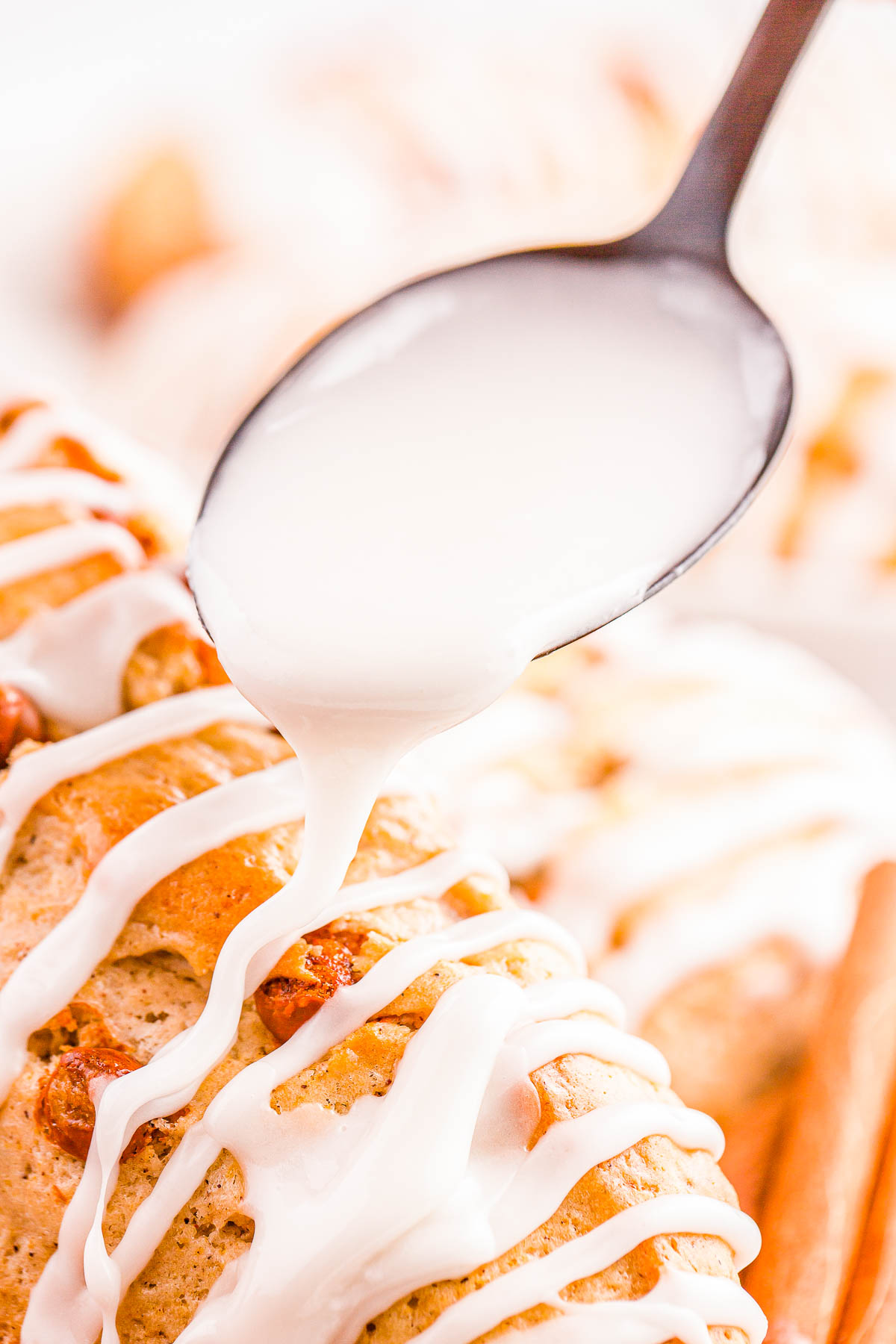 A spoon drizzles white icing over a baked pastry topped with icing and cinnamon sticks nearby.