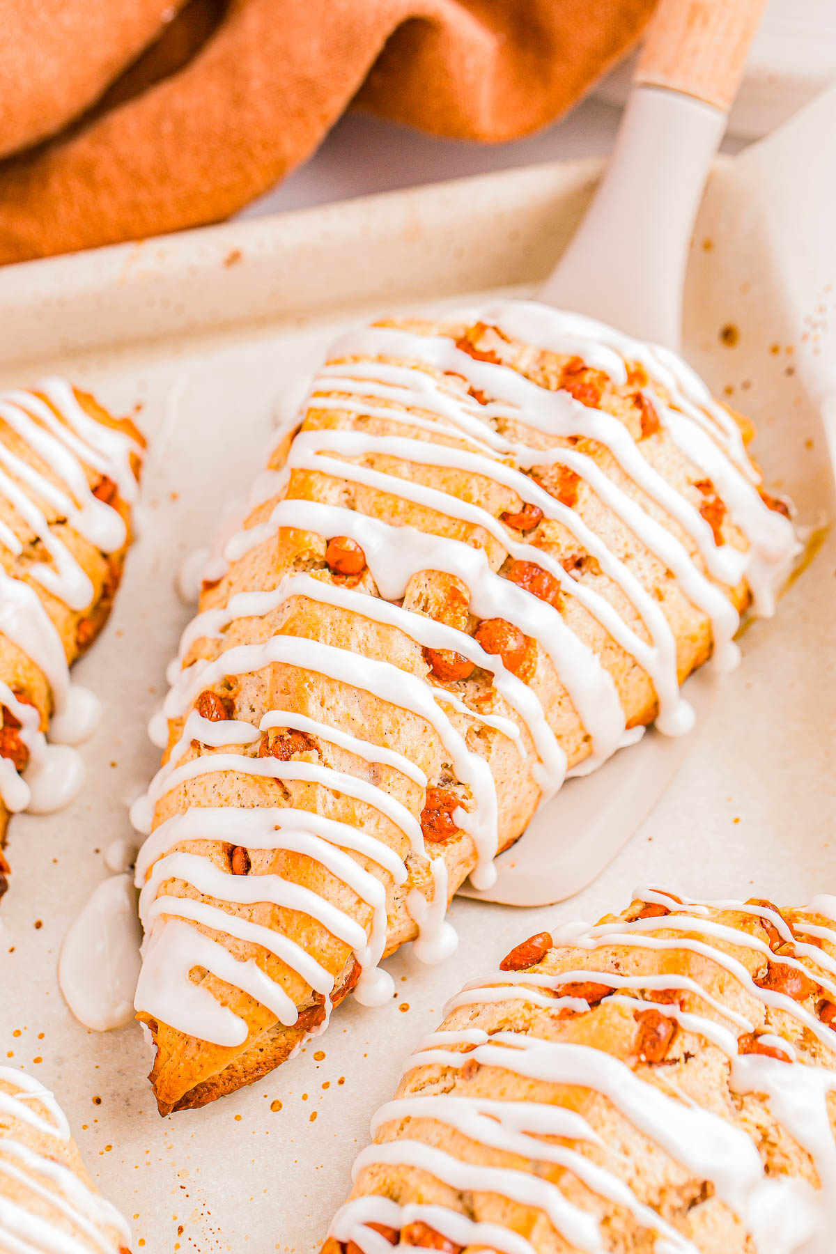 Close-up of glazed scones with pecans on a baking sheet, with a spatula lifting one. A brown cloth is partially visible in the background.