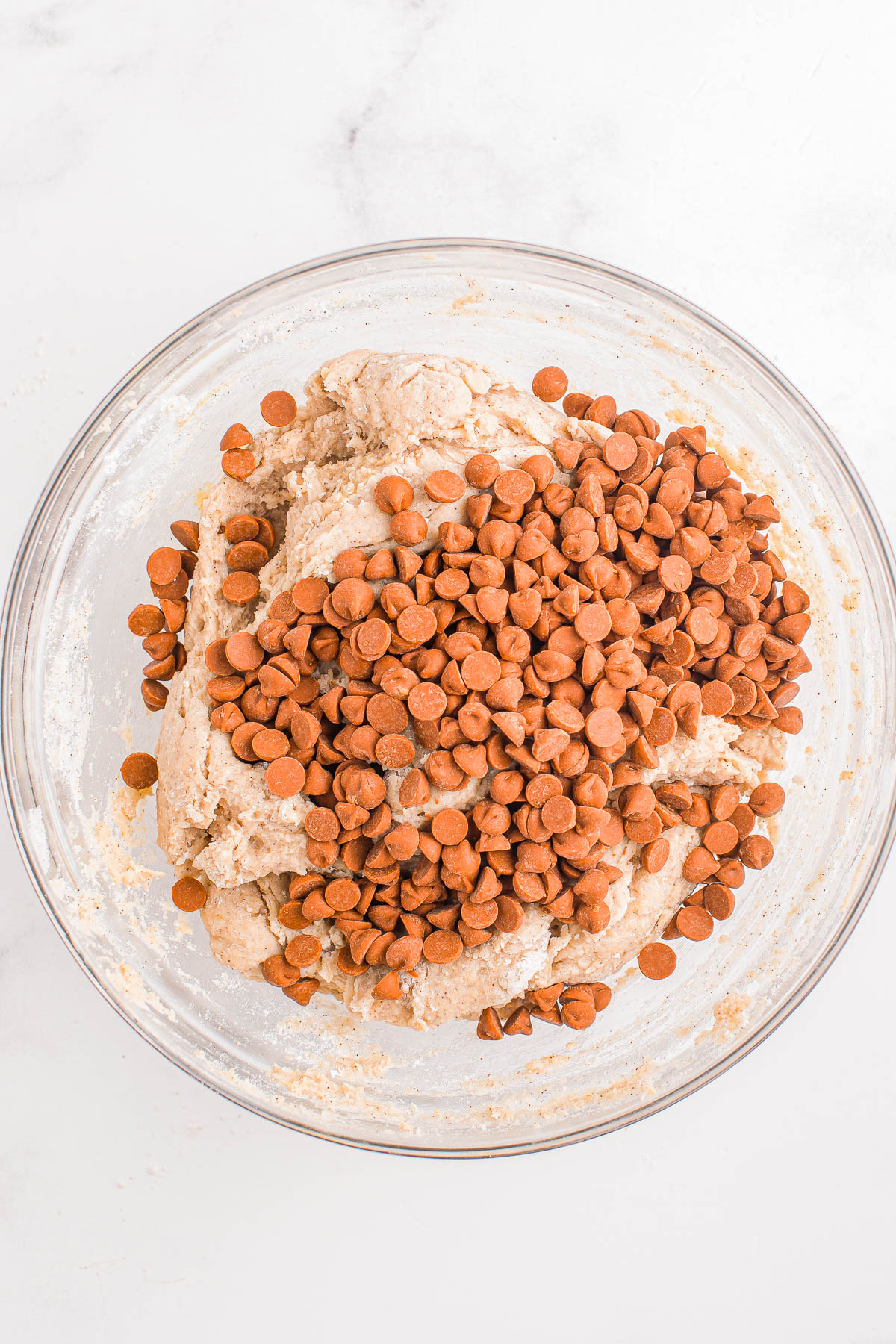 A glass bowl filled with cookie dough topped with chocolate chips on a white marble surface.