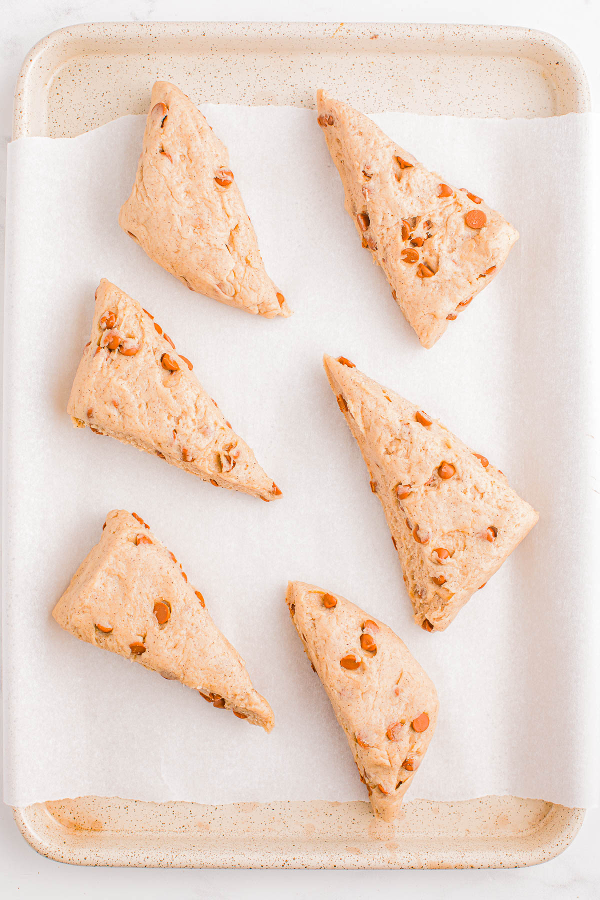 Six triangular scones with visible chocolate chips are arranged on a parchment-lined baking sheet.