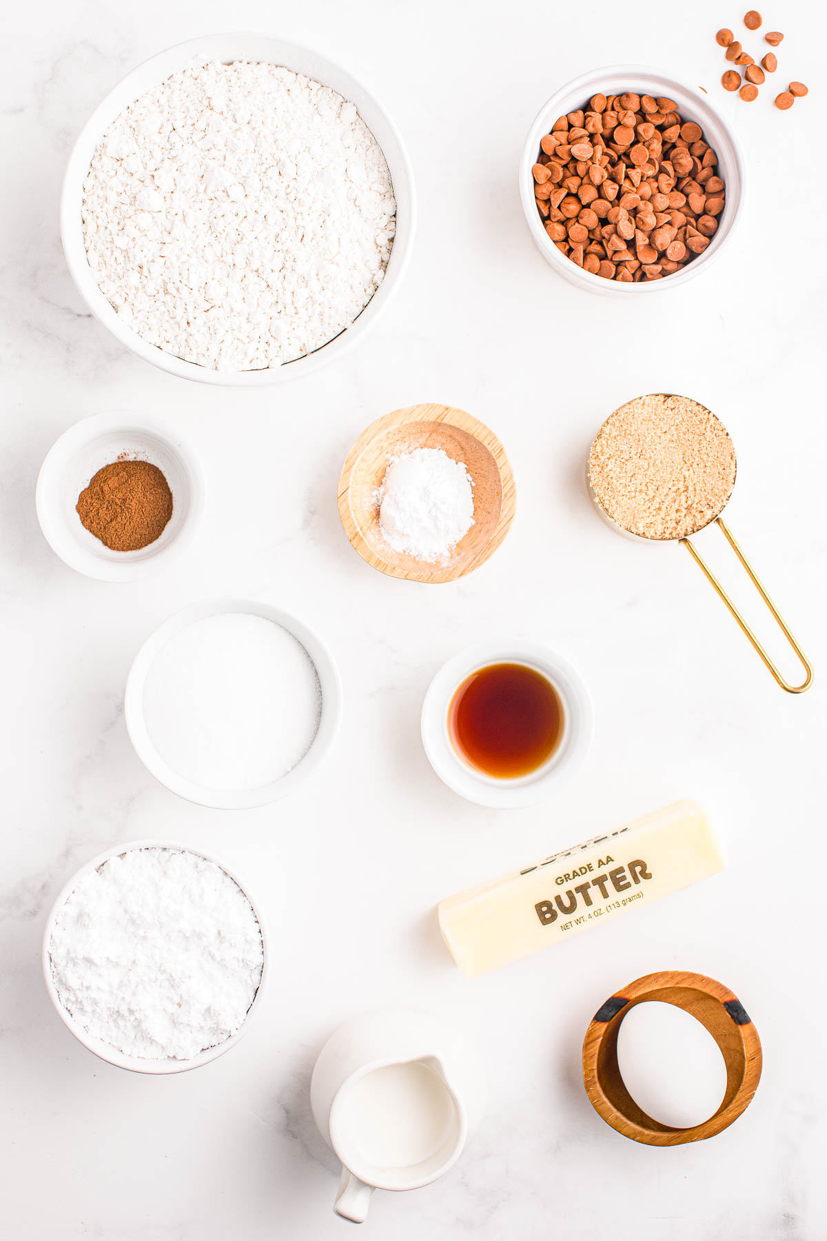 Baking ingredients including flour, cinnamon, sugar, brown sugar, salt, baking powder, vanilla extract, butter, egg, milk, and peanut butter chips are arranged on a white surface.