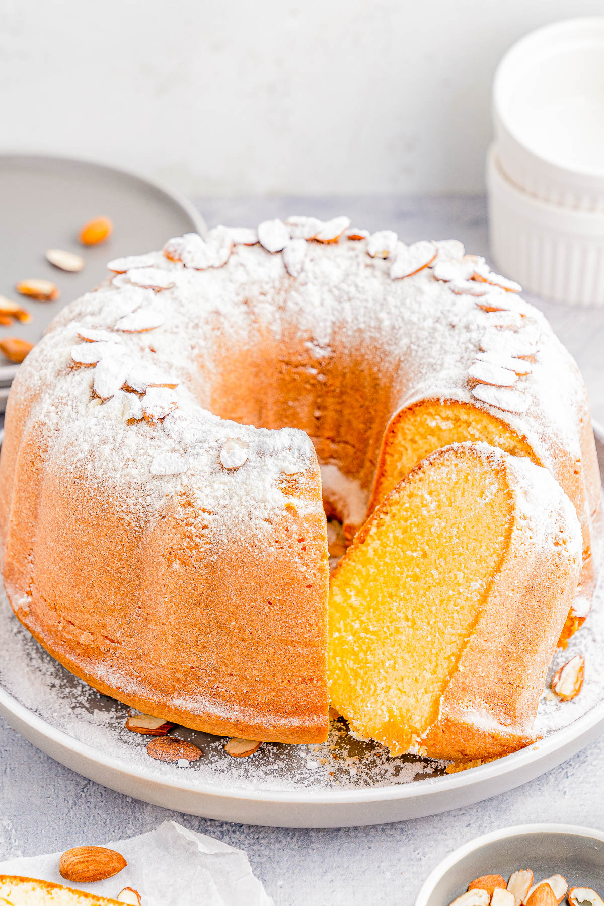 A Bundt cake on a plate, dusted with powdered sugar and topped with sliced almonds. A slice is cut from the cake. Plates and dishes are in the background.