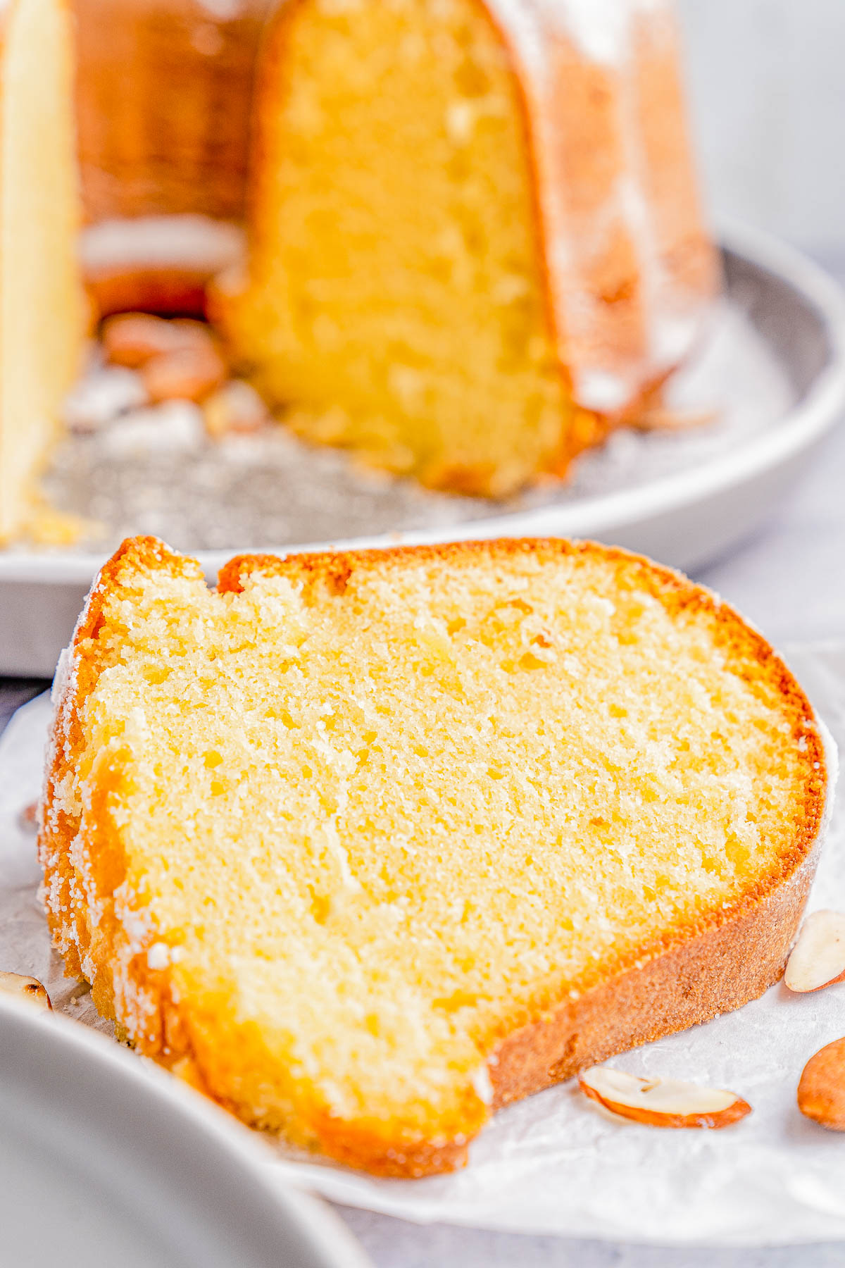 A close-up of a slice of a Bundt cake on a plate, with a dusting of powdered sugar and almond slices nearby.