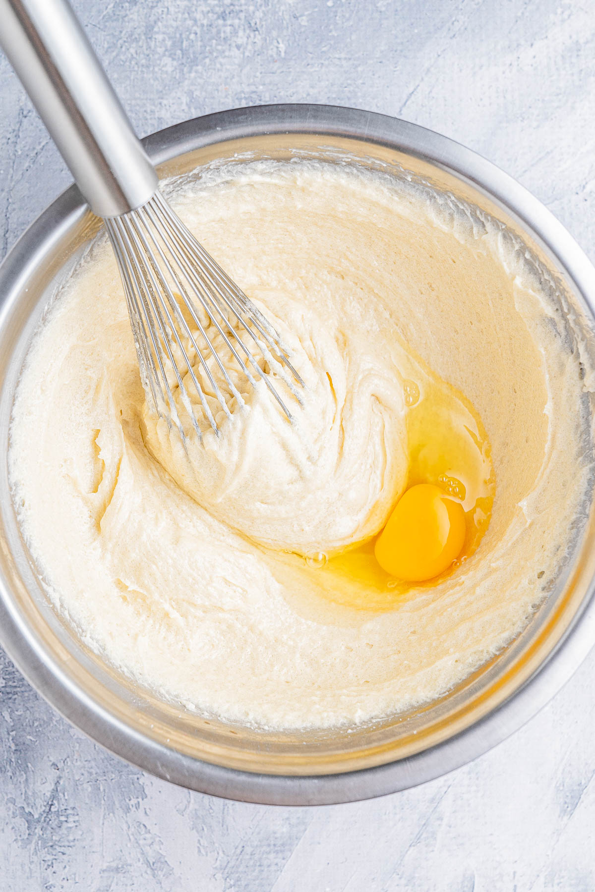 A whisk mixing a yellow egg into a creamy batter in a stainless steel bowl on a textured surface.