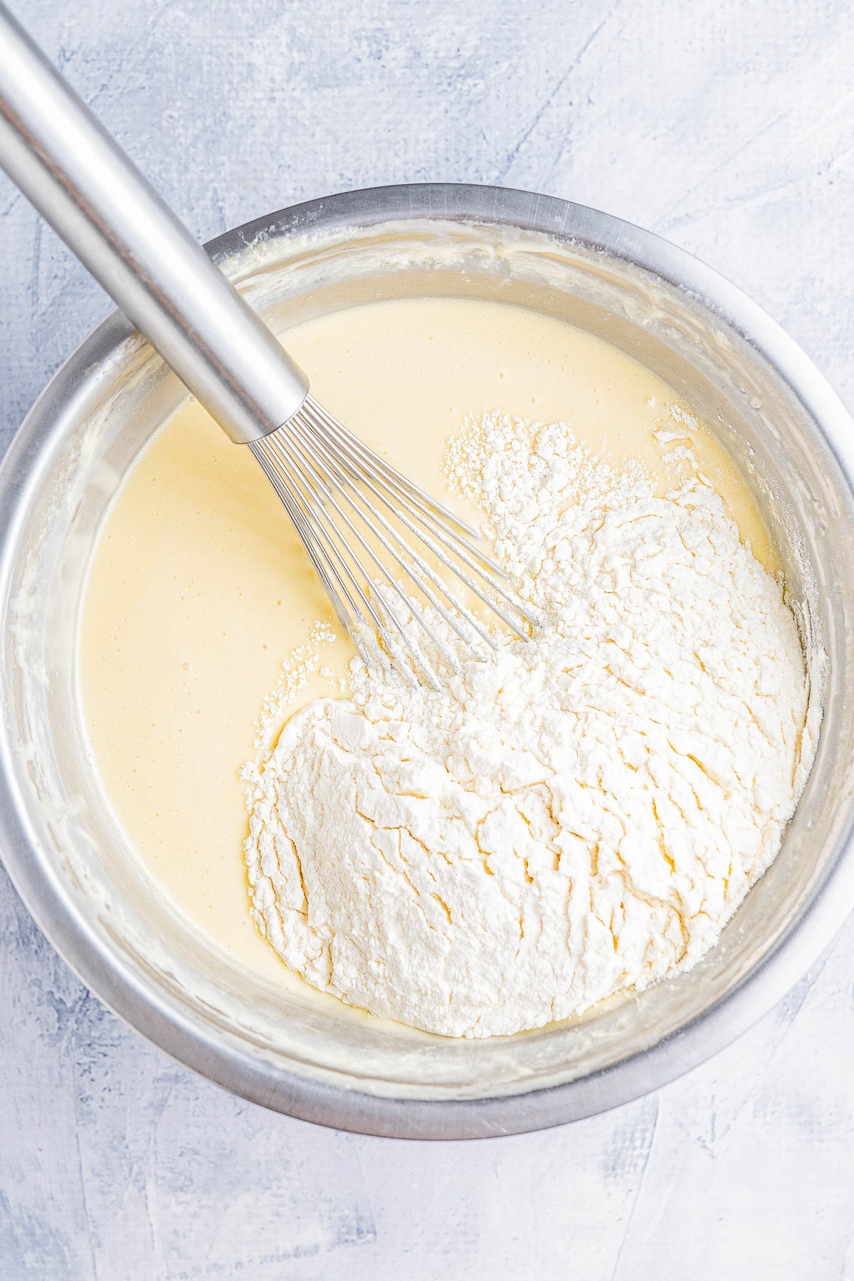 A metal bowl with flour being whisked into a creamy batter using a metal whisk, placed on a light gray textured surface.