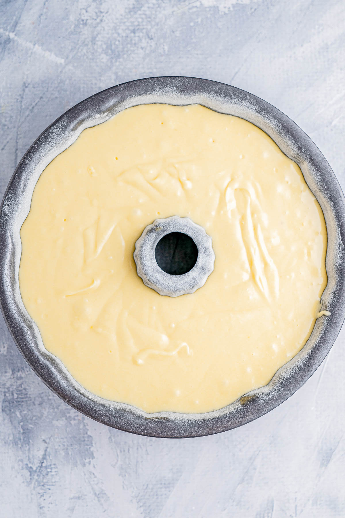 A round, gray bundt pan filled with yellow cake batter, ready for baking, on a light textured surface.