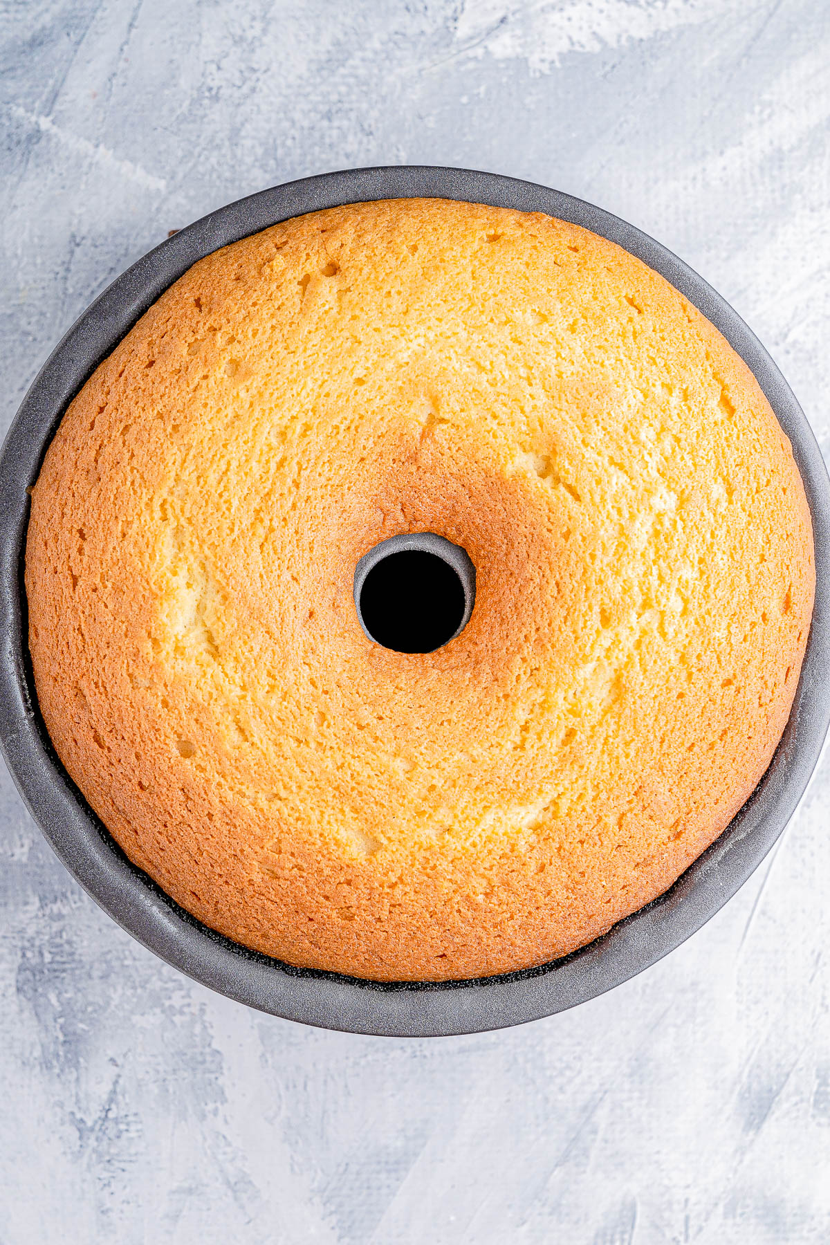 A freshly baked round sponge cake in a bundt pan on a textured gray surface.