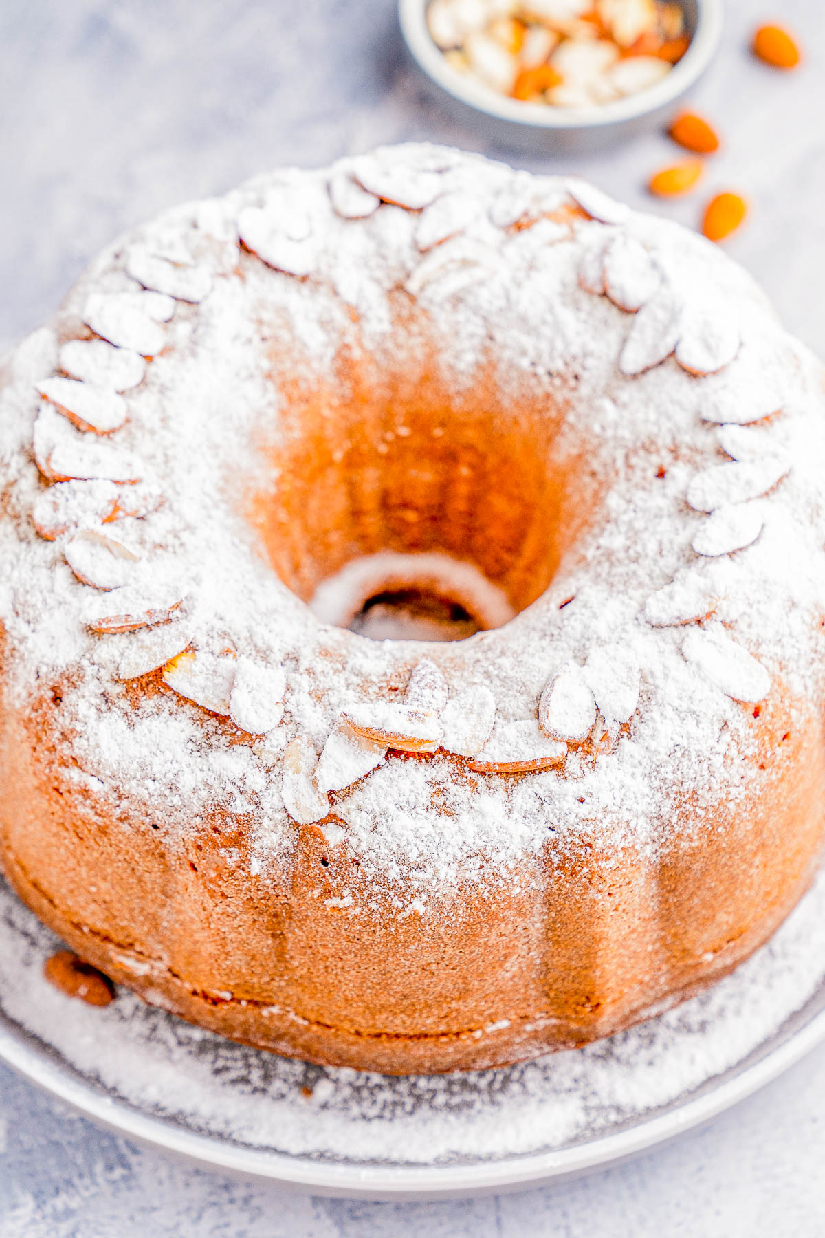 Bundt cake topped with powdered sugar and almond slices on a plate, with a small bowl of almond slices in the background.